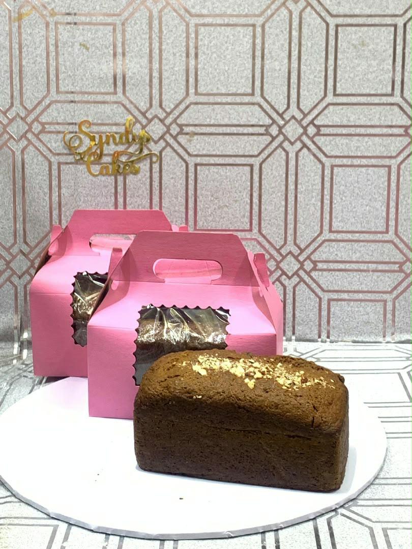 Rectangular chocolate cake loaf with gold flakes on a white board, with pink cake boxes behind it.