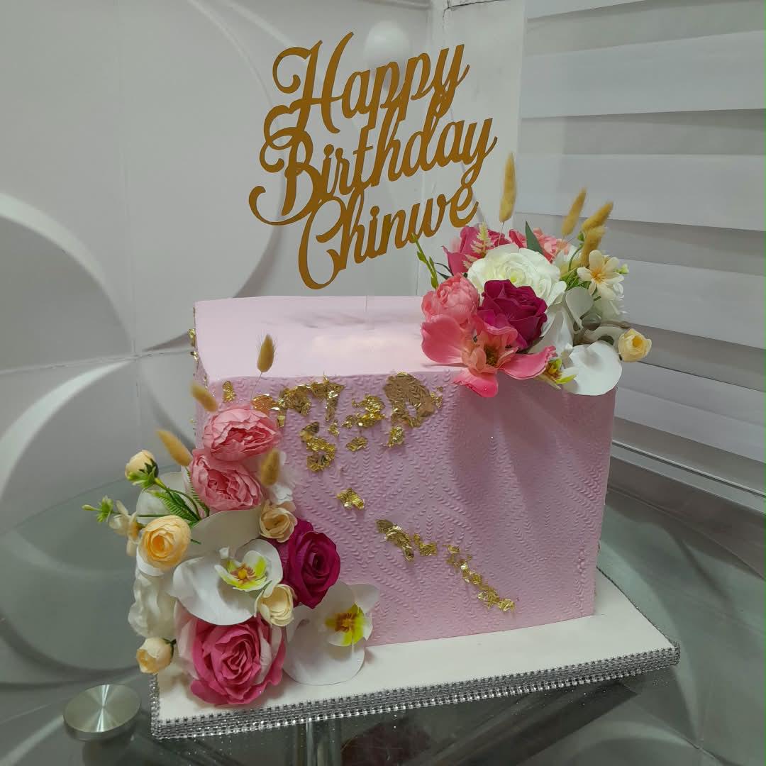 Pink birthday cake with cream flowers and a gold celebration topper.