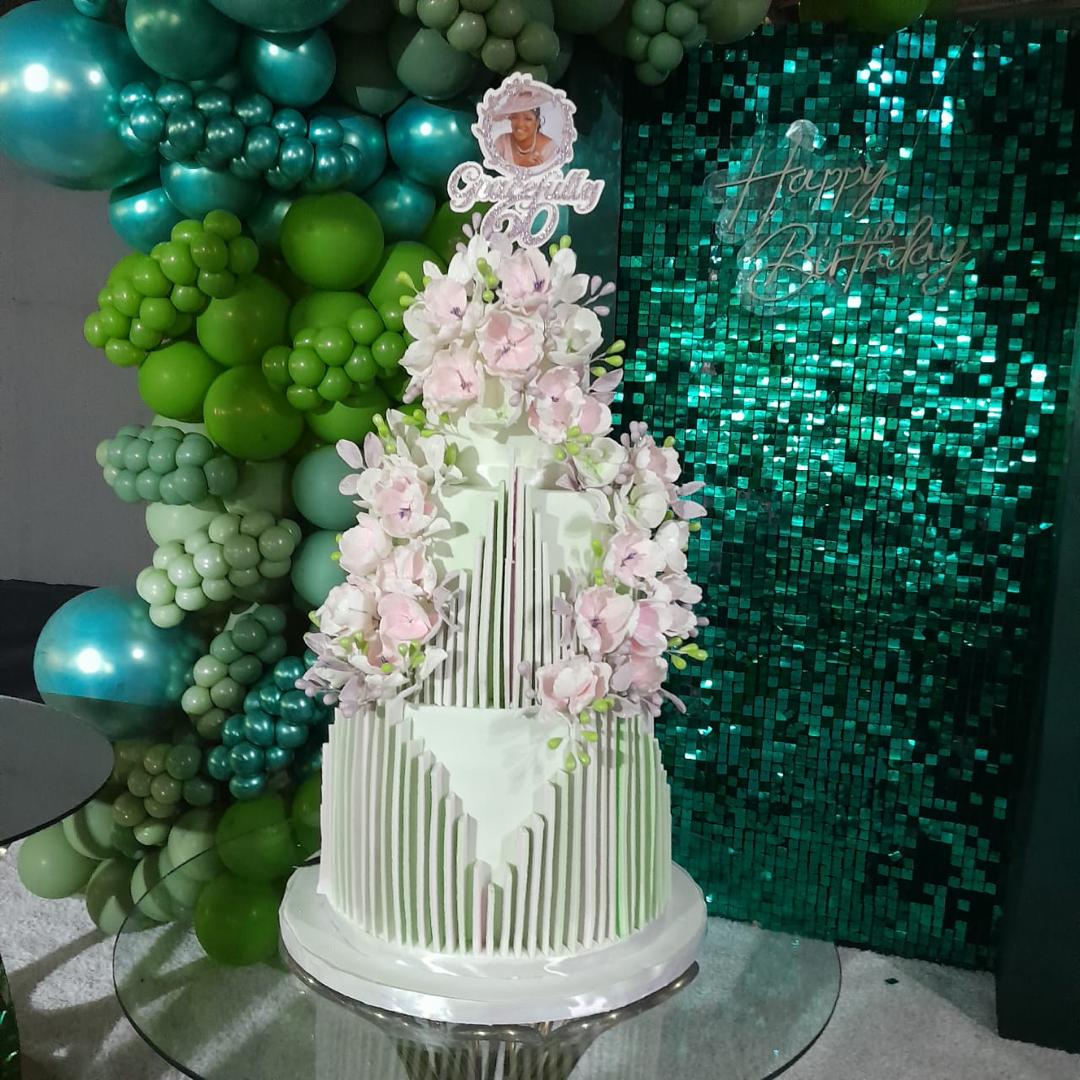 Tall white tiered cake with cascading flowers against a teal balloon backdrop.