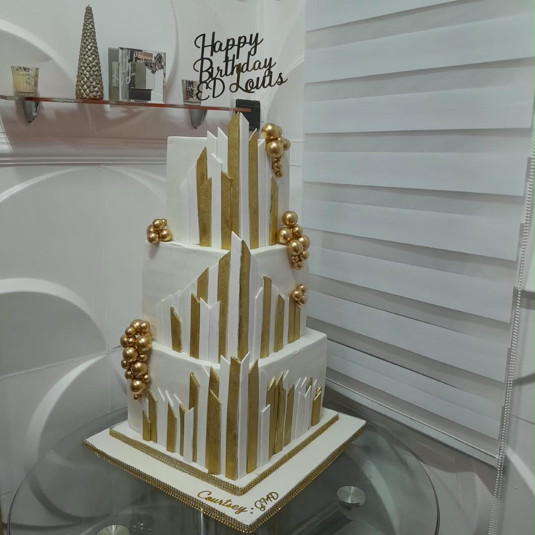 Tall white tiered cake with gold vertical accents and a celebration topper.