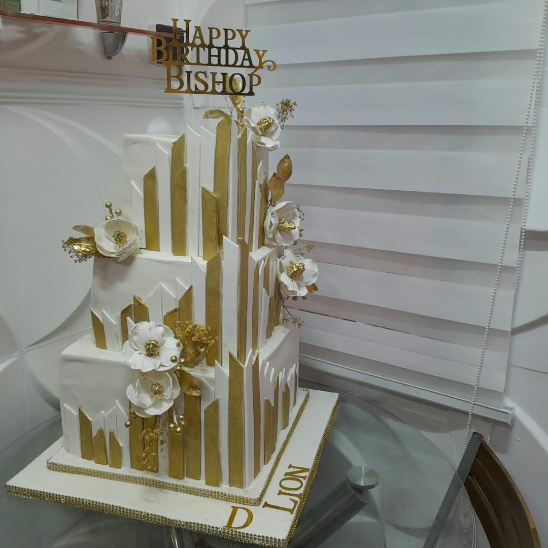 White-and-gold tiered cake with flowers and a Happy Birthday Bishop topper.